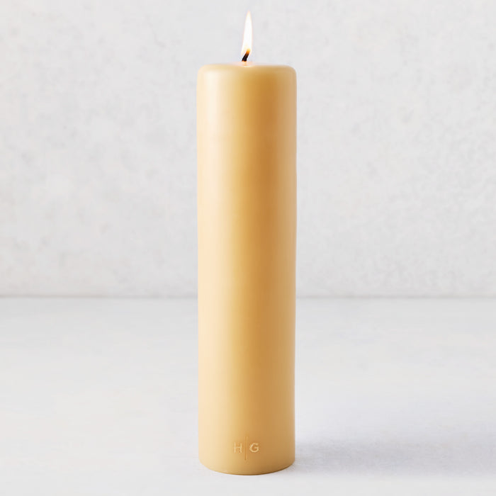 3x12 inch amber unscented paraffin pillar candle with a lit wick and embossed gold HG logo on a light background.