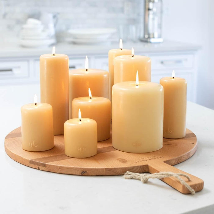 Amber unscented paraffin pillar candles in various sizes, hand-embossed with a gold HG logo, on a wooden board.