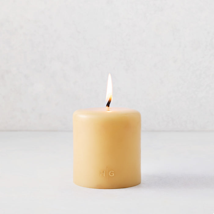 4x4 inch amber pillar candle with a lit cotton wick and embossed gold HG logo on a light background.