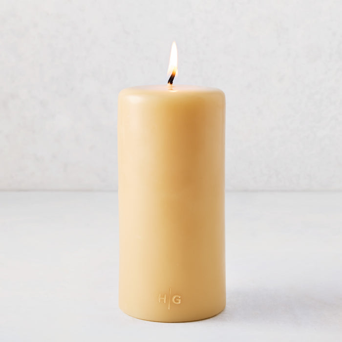 4x8 inch amber unscented pillar candle with a lit cotton wick and gold embossed HG logo on a light background.