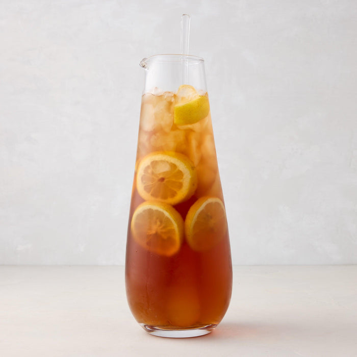 Clear glass cocktail jug with stirrer, filled with iced tea and lemon slices.