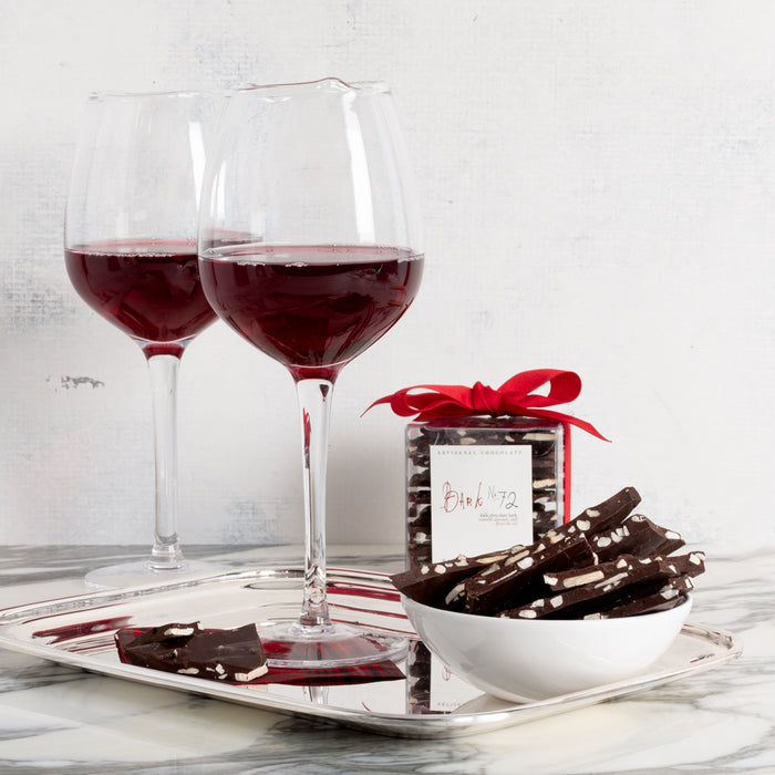 Bark 72 Dark Chocolate Almond Bark in a clear box with red ribbon, a white bowl of bark pieces, and two red wine glasses on a silver tray.