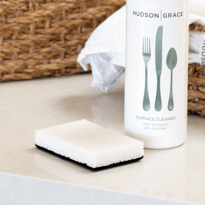 White and black rectangular kitchen sponge with a non-scratch scourer, next to a Hudson Grace cleaner bottle.