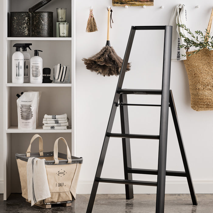 Black "Bon Appetit" linen hand towel with green sprigs in a woven basket, next to a black step ladder.