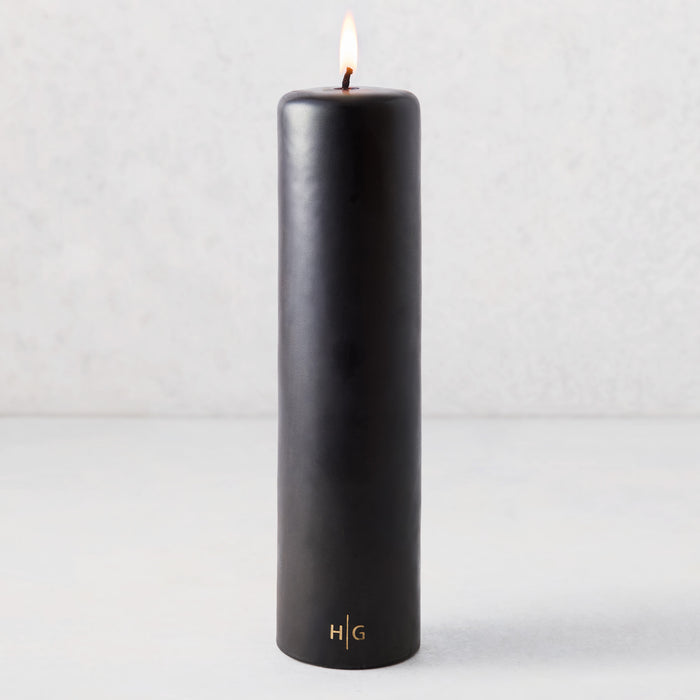 3x12 inch black unscented pillar candle with a gold HG logo and a lit cotton wick.