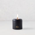 3x3 inch black hand-dipped pillar candle with a burning wick and gold embossed HG logo on a white surface.