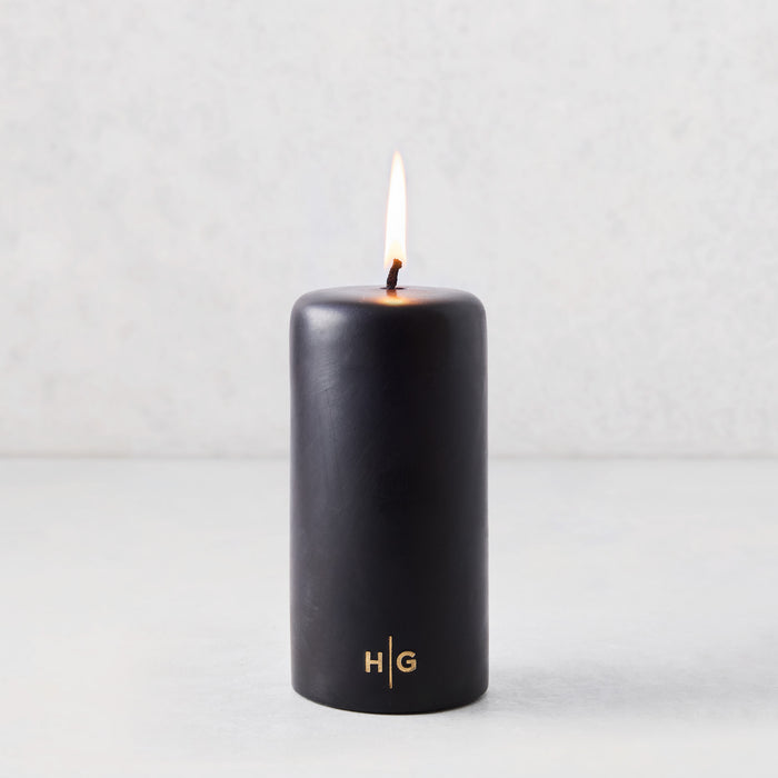 3x6 inch black unscented pillar candle with a burning wick and gold HG logo on a light grey background.
