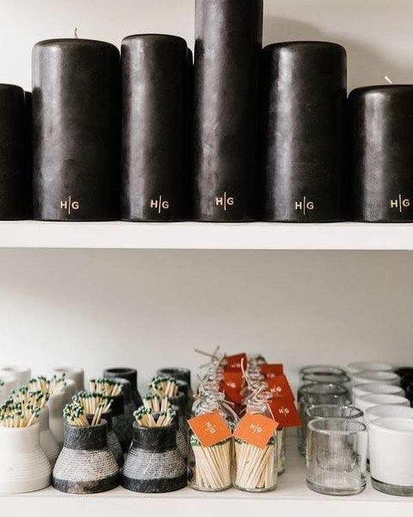 HG Black Unscented Pillar Candle, 3"x6", with gold HG logo, on a white shelf.