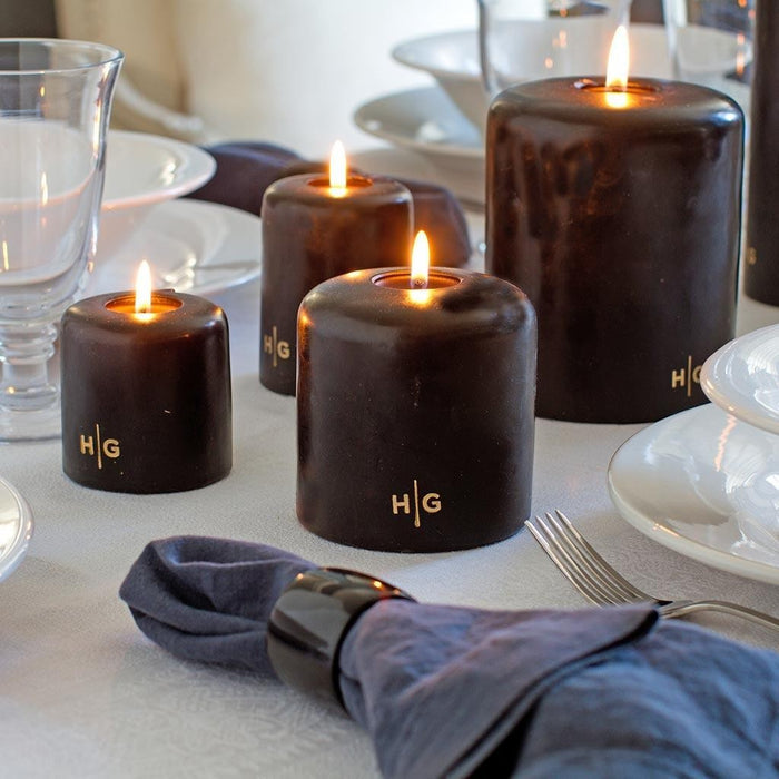 HG Black Unscented Pillar Candles, 3"x6" and smaller, with gold HG logo, lit on a dining table.
