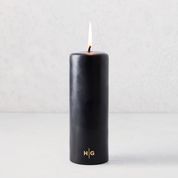 3x8 inch black unscented pillar candle with a gold HG logo and lit cotton wick.