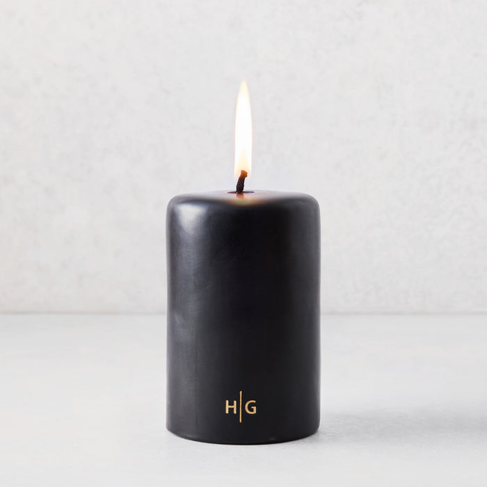 4x6 inch black unscented pillar candle with a burning wick and gold HG logo on a light background.