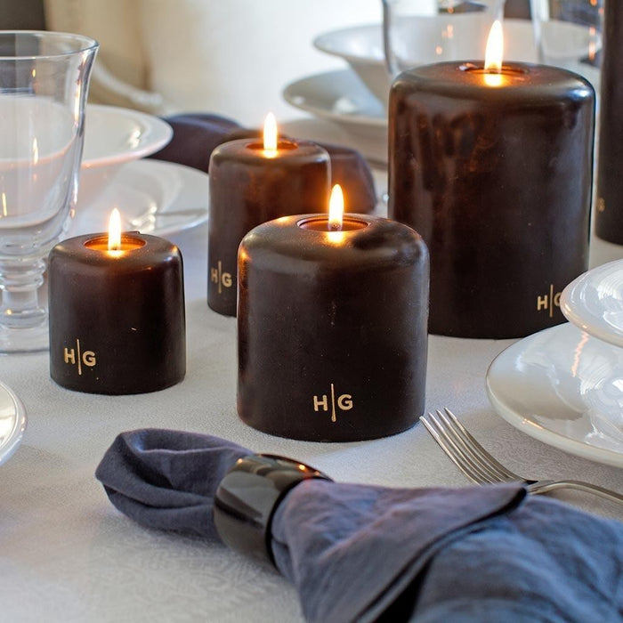 HG Black Unscented Pillar Candles, 4"x6" and smaller, with gold HG logo, on a white dining table.