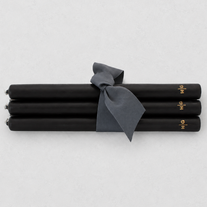 Four black taper candles tied together with a dark gray ribbon, featuring small gold lettering near the ends, on a light gray background.