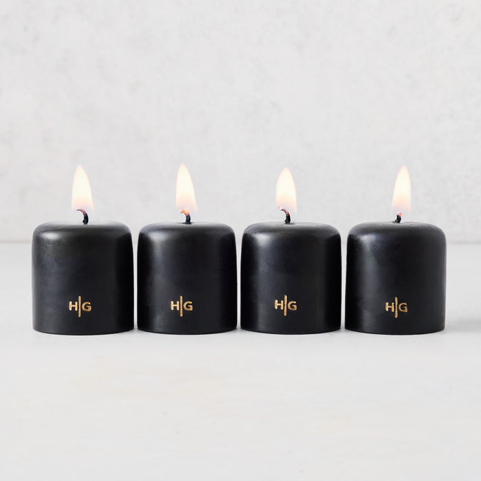 Set of four 2-inch black unscented votive candles with lit wicks and gold embossed HG logo on a light background.