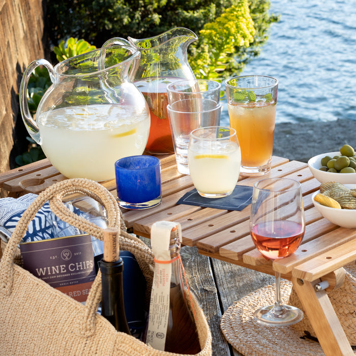 Blue votive glass candle holder on a wooden picnic table with drinks and snacks by water.