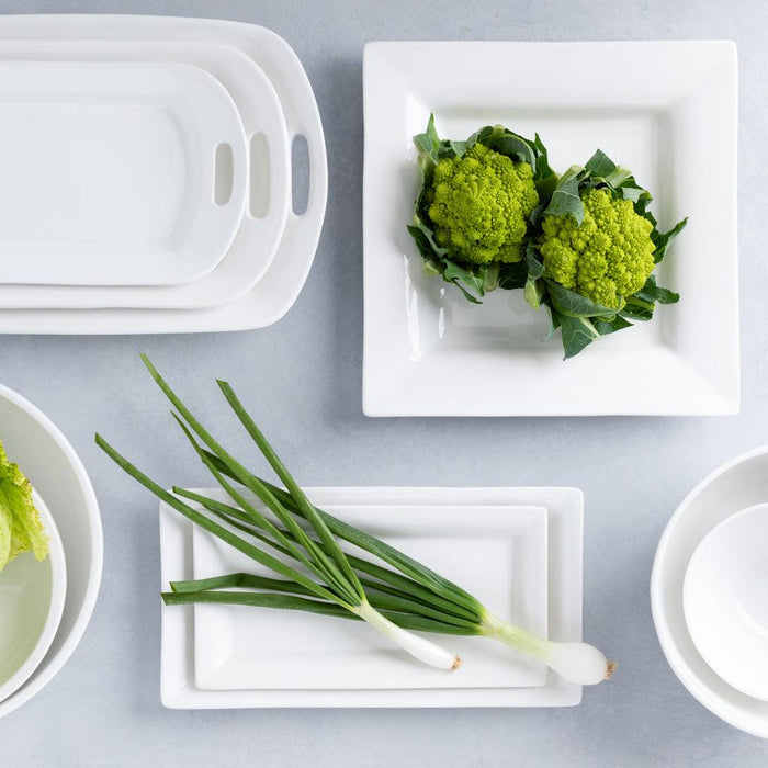 White ceramic handled serving platter, rectangular, with other white dishes and green vegetables.
