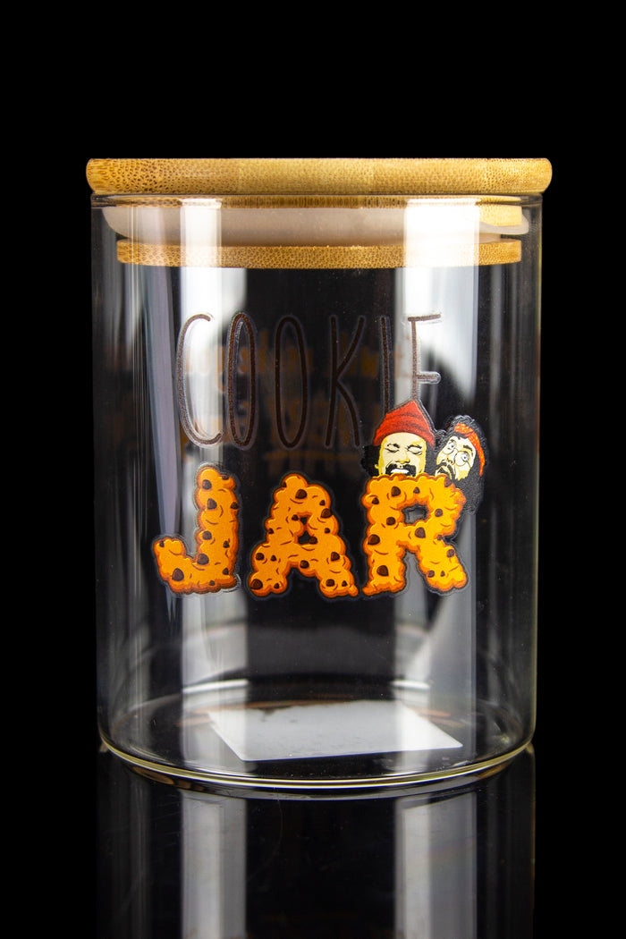 Glass “Cookie Jar” storage container with cartoon cannabis-themed graphics and a bamboo lid, displayed on a reflective black surface.