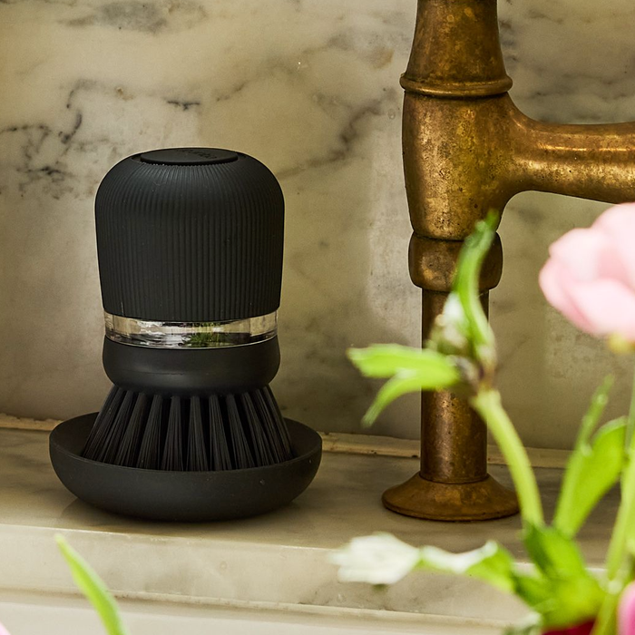Dark grey soap dispensing dish brush with polyester bristles and clear reservoir, on a matching stand.
