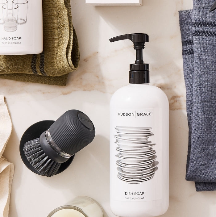 Dark grey soap dispensing dish brush with stand, next to a white dish soap bottle.