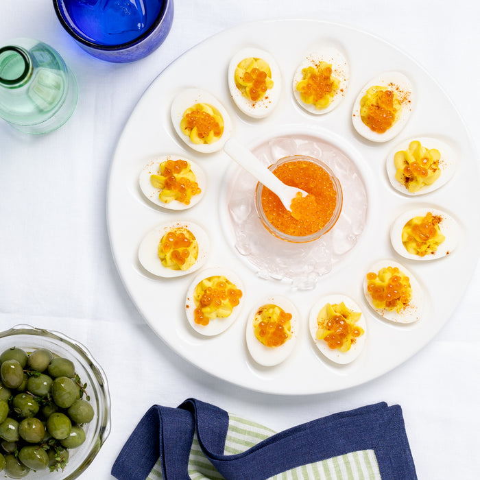 White ceramic 11"Dia Deviled Egg Serving Platter with 12 deviled eggs, a center dip, and blue glass.
