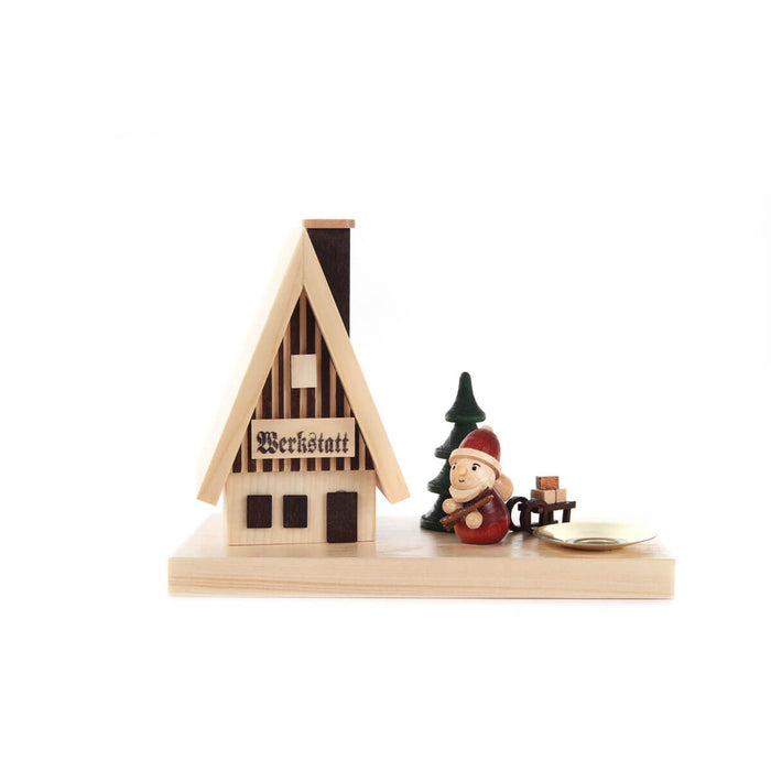 This German Smoker House - Workshop is a charming piece for your holiday home decor. The design features a wooden workshop – Werkstatt - with a little man and a sled with wooden blocks and a candle holder, set against a green tree. Perfect for adding some traditional German style to your celebration.