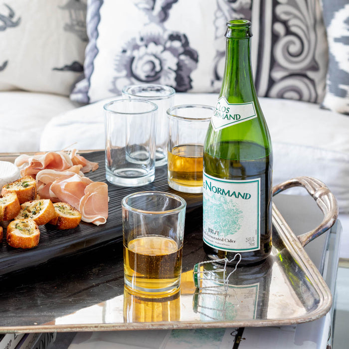 Clear, simple 8.5 oz Everyday Wine Glasses, some filled with amber liquid, next to a green cider bottle and appetizers.