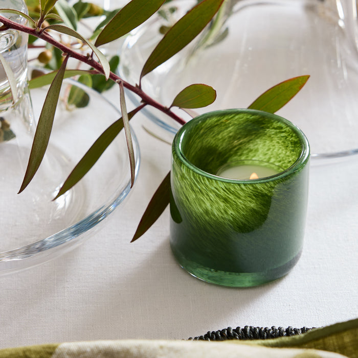 Hand-formed, mouth-blown green votive glass candle holder with a lit candle on a white tablecloth.