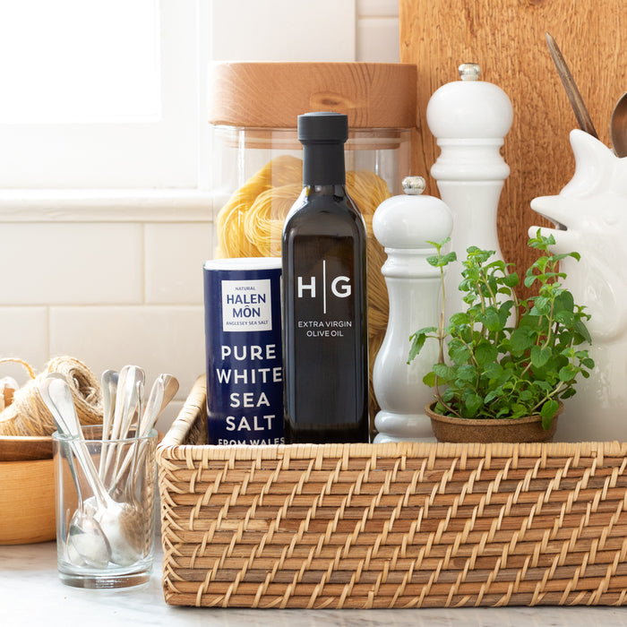Halen Mon Pure White Sea Salt in blue container, black olive oil bottle, and white shakers in a woven basket.