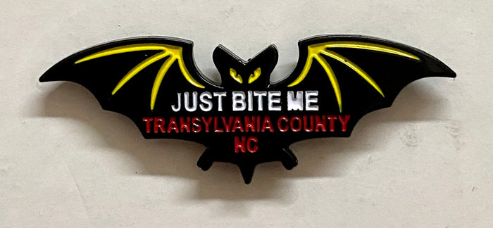 A metal black bat hat/lapel pin with the text 'JUST BITE ME, TRANSYLVANIA COUNTY, NC' on it.