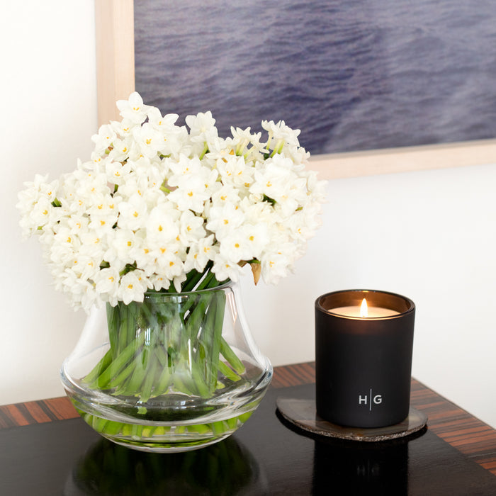 Hudson Grace Bleecker scented candle in a matte black glass vessel with HG logo, next to white flowers.