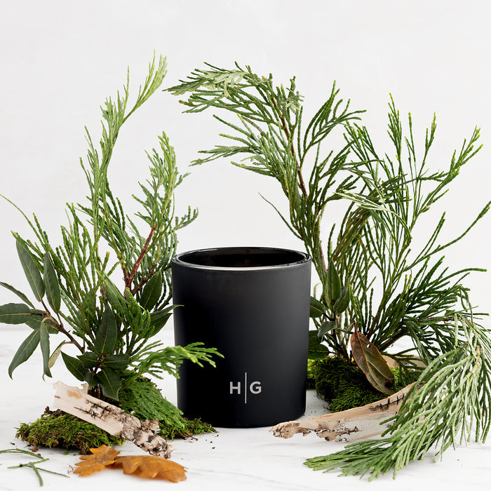 Hudson Grace Bleecker Scented Candle, black matte glass vessel with HG logo, surrounded by green foliage and birch bark.