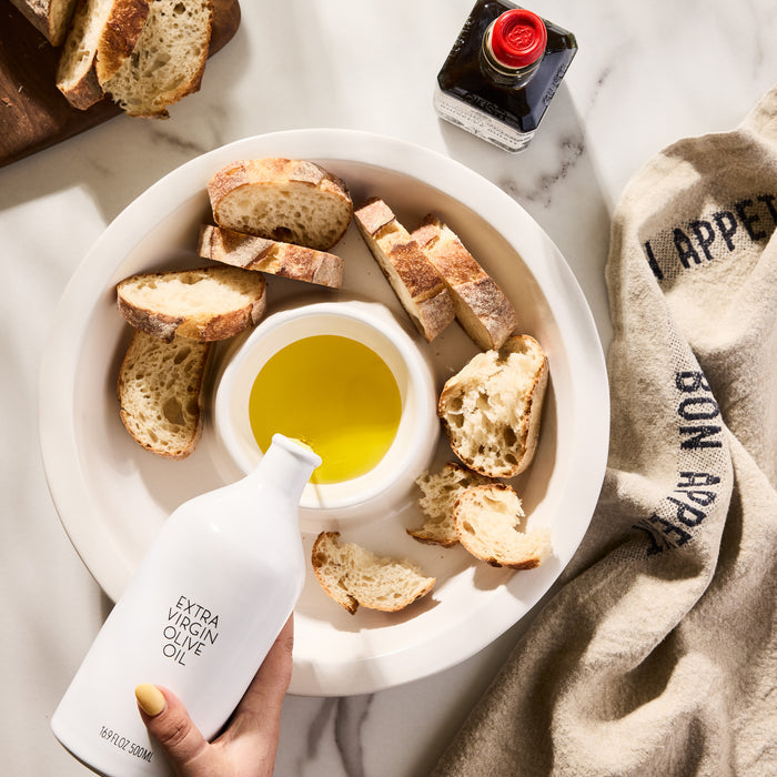 White ceramic Hudson Grace Extra Virgin Olive Oil bottle pouring into a dipping dish with bread slices.