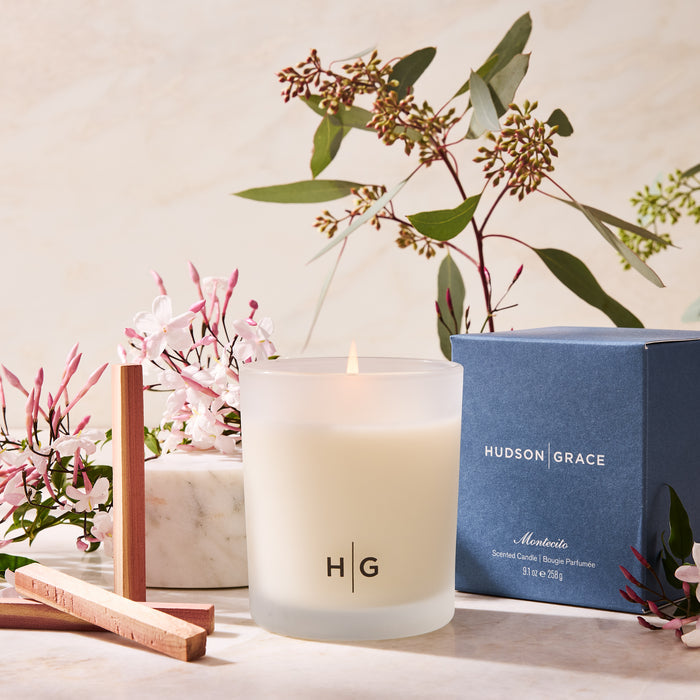 Hudson Grace Montecito scented candle in a frosted glass vessel with a burning wick, next to its blue box.