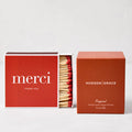 Hudson Grace Original Scented Candle in a rust-colored box next to a red "merci" matchbox with red-tipped matches.