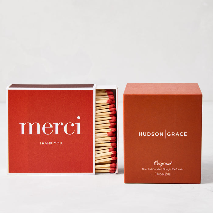Hudson Grace Original Scented Candle in a rust-colored box next to a red "merci" matchbox with red-tipped matches.