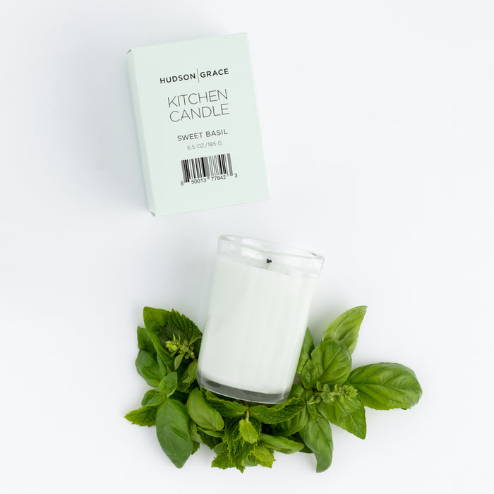 Hudson Grace Sweet Basil Kitchen Candle in a clear glass, surrounded by fresh basil and mint leaves, with its light green box.