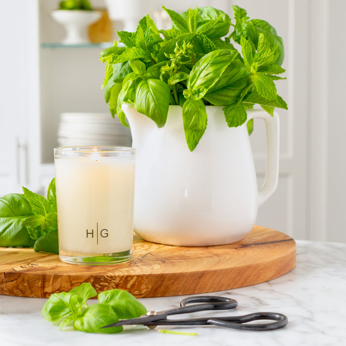 Hudson Grace Sweet Basil Kitchen Candle, lit, in a clear glass jar with "HG" logo, next to a white pitcher of fresh basil and mint on a wooden board.