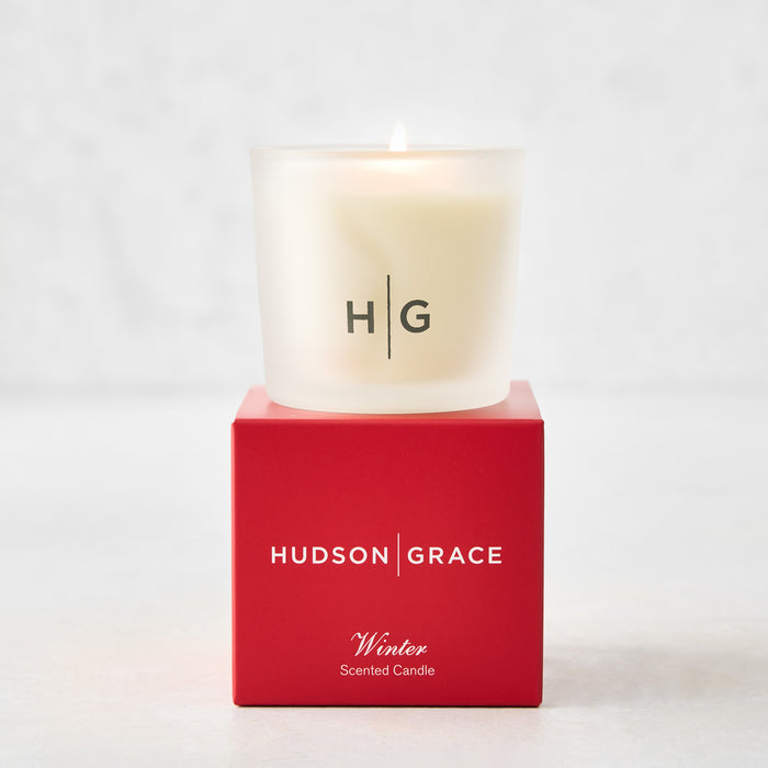 Hudson Grace Winter Scented Votive Candle in a frosted glass vessel, lit, on a red gift box.