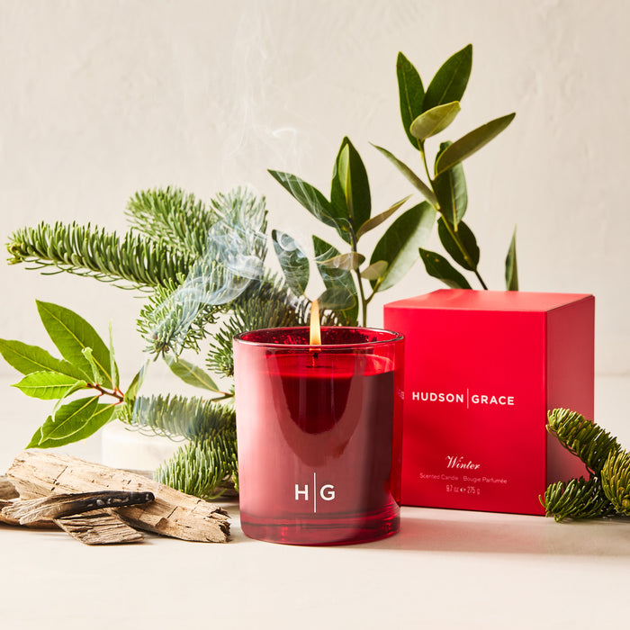 Hudson Grace Winter Scented Votive Candle in a red glass vessel, lit with smoke, next to a red gift box.
