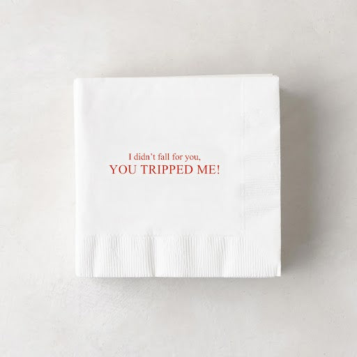 White paper cocktail napkin with "I didn't fall for you, YOU TRIPPED ME!" in red text.