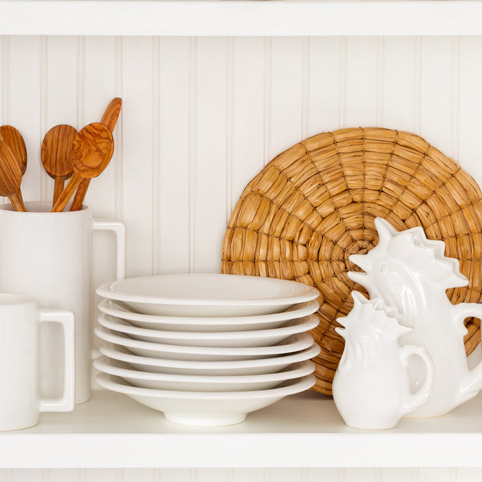 Stack of white ceramic pasta bowls, mugs, and rooster pitchers with wooden spoons and a woven placemat.