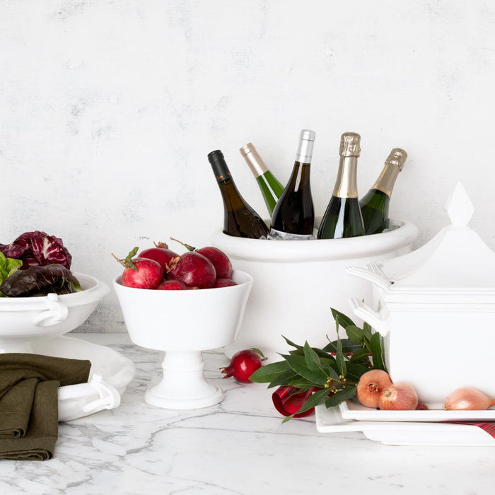 Creamy white Italian ceramic compote bowl with pomegranates, surrounded by other white serveware and wine bottles.