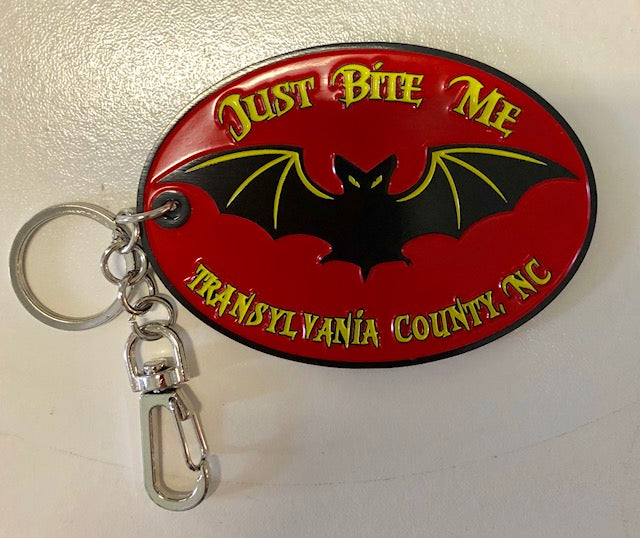 A metal key chain clip with a black bat design and the text 'Just Bite Me, Transylvania County, NC' on a red background.