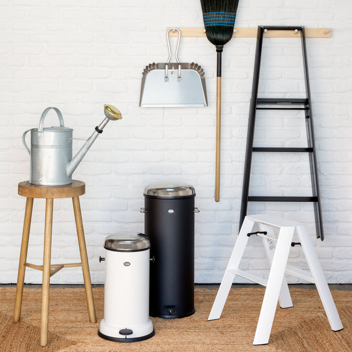 Silver aluminum dust pan with a wooden handle, hanging on a white brick wall.
