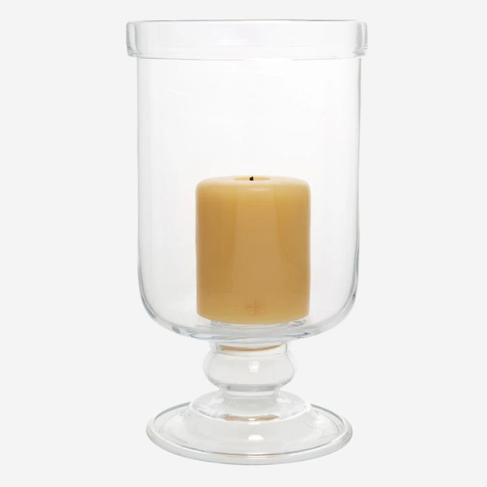 Large clear glass footed chalice hurricane candle holder with a folded edge, holding a beige pillar candle.