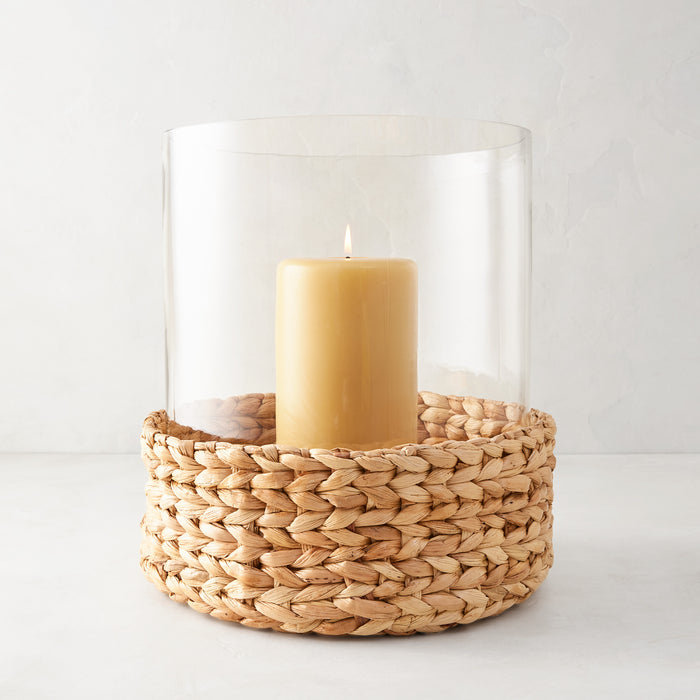 Large glass hurricane with woven water hyacinth base and lit amber candle.