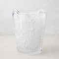 Large clear handblown glass ice bucket with two handles, filled with ice.