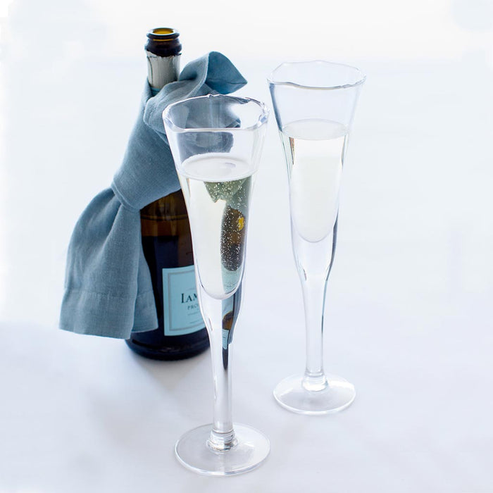Two clear, thick, scissor-cut glass champagne flutes with a rippled rim, filled with bubbly, next to a champagne bottle.