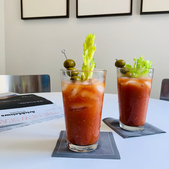 Two tall clear glass tumblers filled with Bloody Mary cocktails, garnished with celery and green olives.