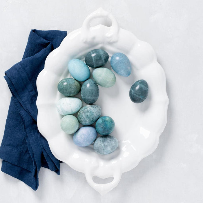 White ceramic oval serving platter with handles, holding blue and green dyed eggs, next to a navy linen napkin.